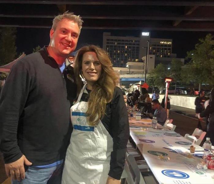 Image shows two people posing in front of a very long table, one wears an apron.