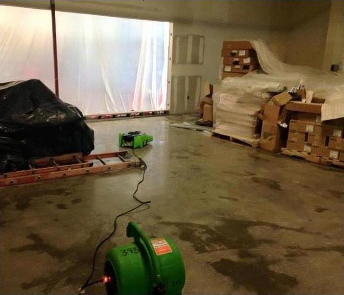 Image shows concrete warehouse floor with water and green fans with building materials stacked up.